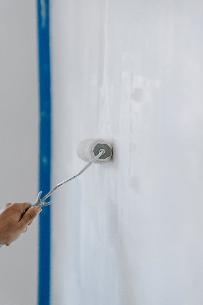 A close-up of a person using a paint roller to apply white paint on an indoor wall.